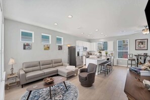 A modern living room with a sofa, armchairs, and a coffee table. A kitchen with white cabinets and a dining area with stools is visible.