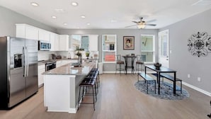 A modern kitchen with a central island, stainless steel appliances, and a dining area with a ceiling fan. - Magnolia Haus at Solms Landing (New Braunfels)