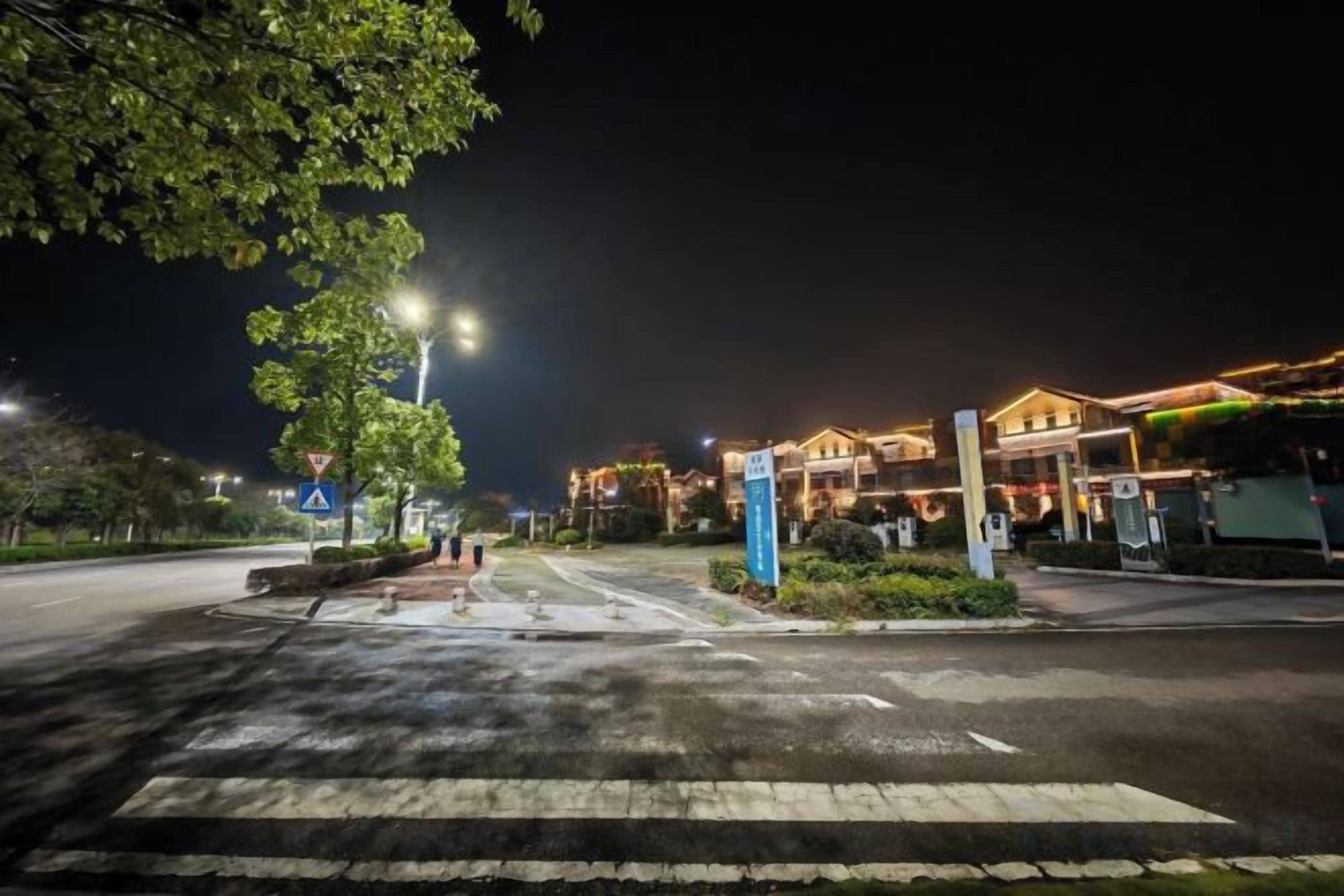 Straßenansicht bei Nacht mit Fußgängerüberweg, Straßenlaternen und einem Gebäude mit beleuchteten Fenstern.