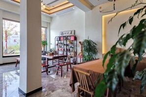 A spacious dining area with a high ceiling, a central table, and chairs. There is a bookshelf with various items and a large potted plant. - Wuyishan Hike Tea House Inn (Wuyishan)