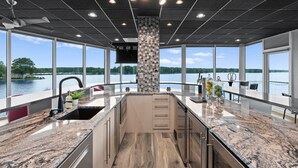 A modern kitchen with a large island, a central column, and large windows overlooking a body of water. - The Glass Mansion w/Optional Boat Rental (Terrell)