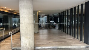 Un vestíbulo moderno con suelo de madera, un pilar de hormigón y puertas de cristal que dan a un estacionamiento con coches y una motocicleta.