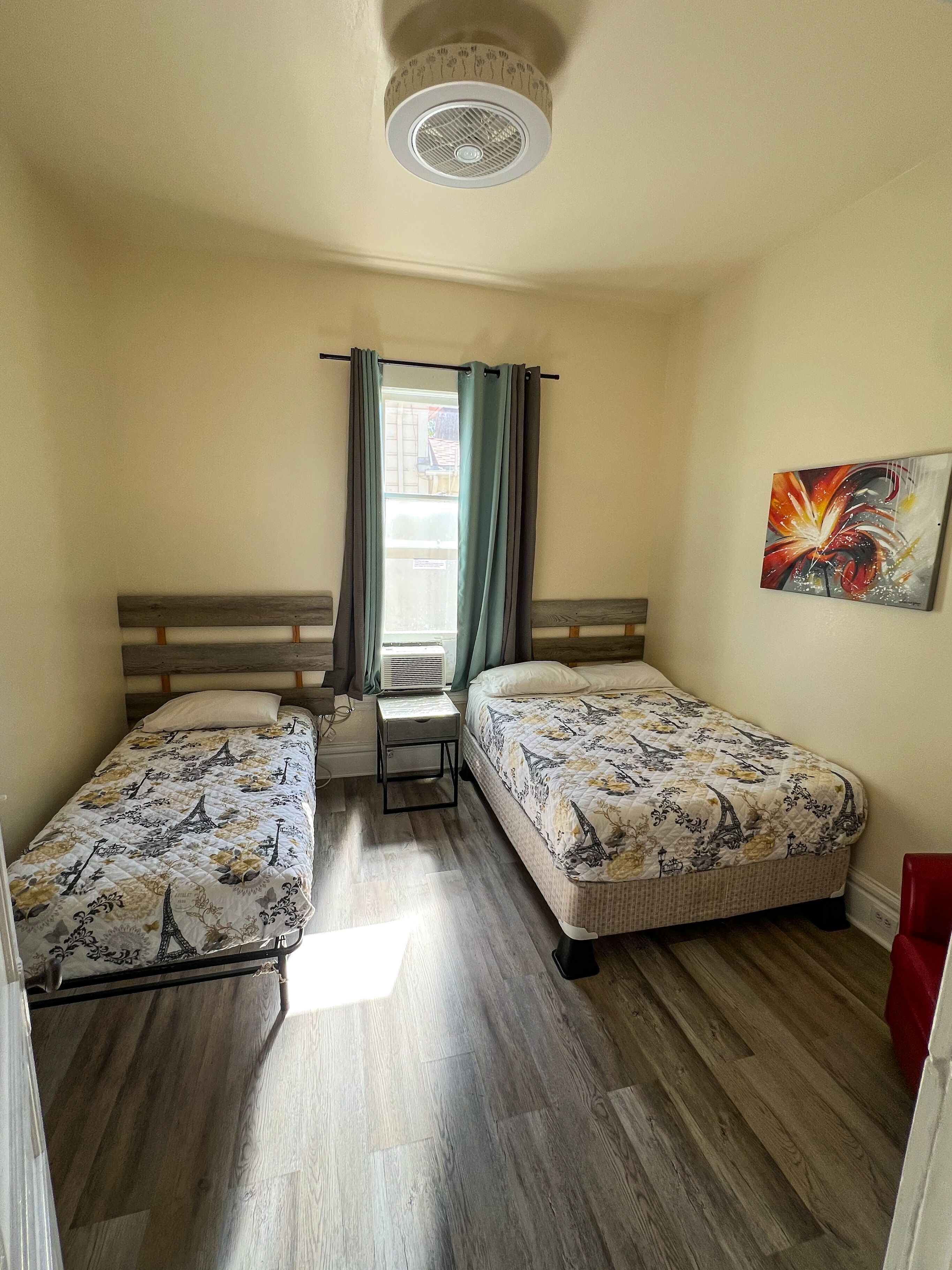 Una habitación pequeña y luminosa con dos camas individuales, una ventana con cortinas, un ventilador de techo y un cuadro en la pared.