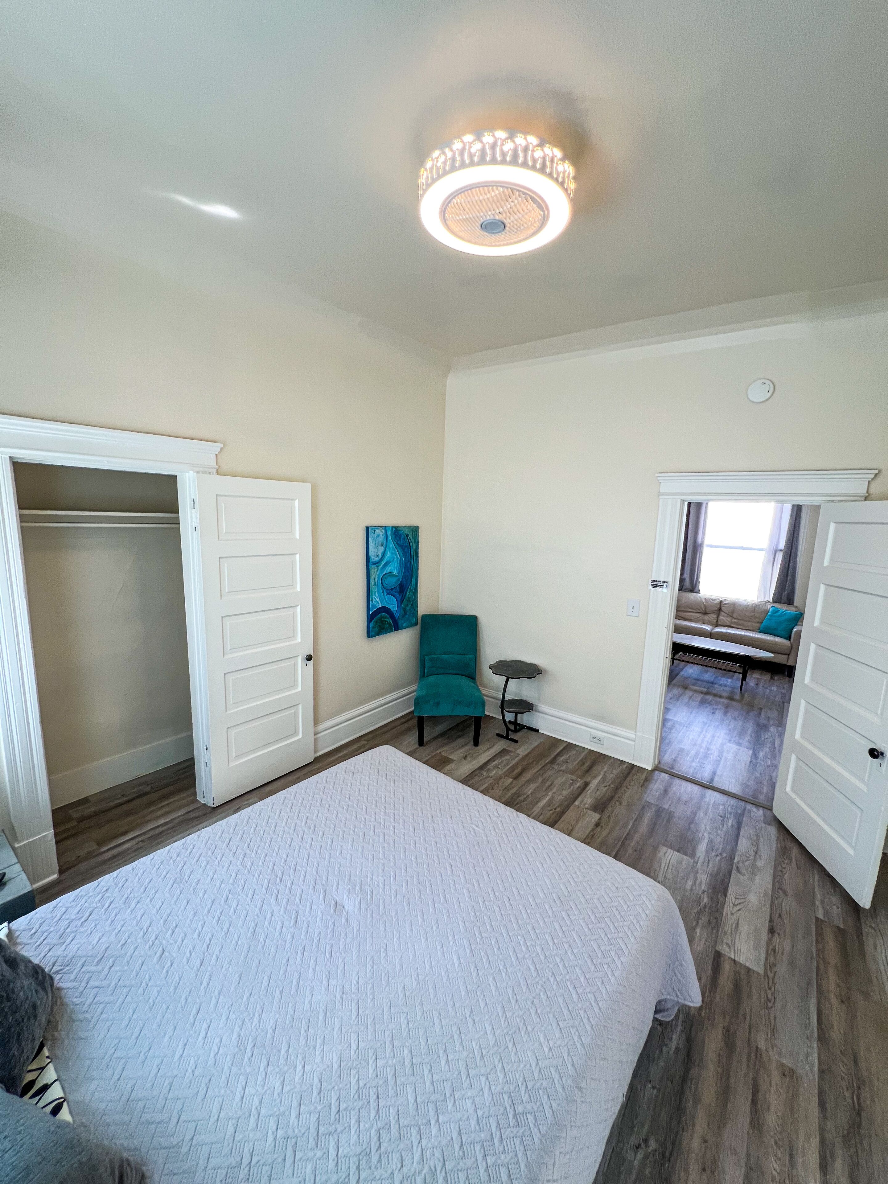 Een ruime slaapkamer met een groot bed, een blauwgroene fauteuil en uitzicht op een andere kamer.