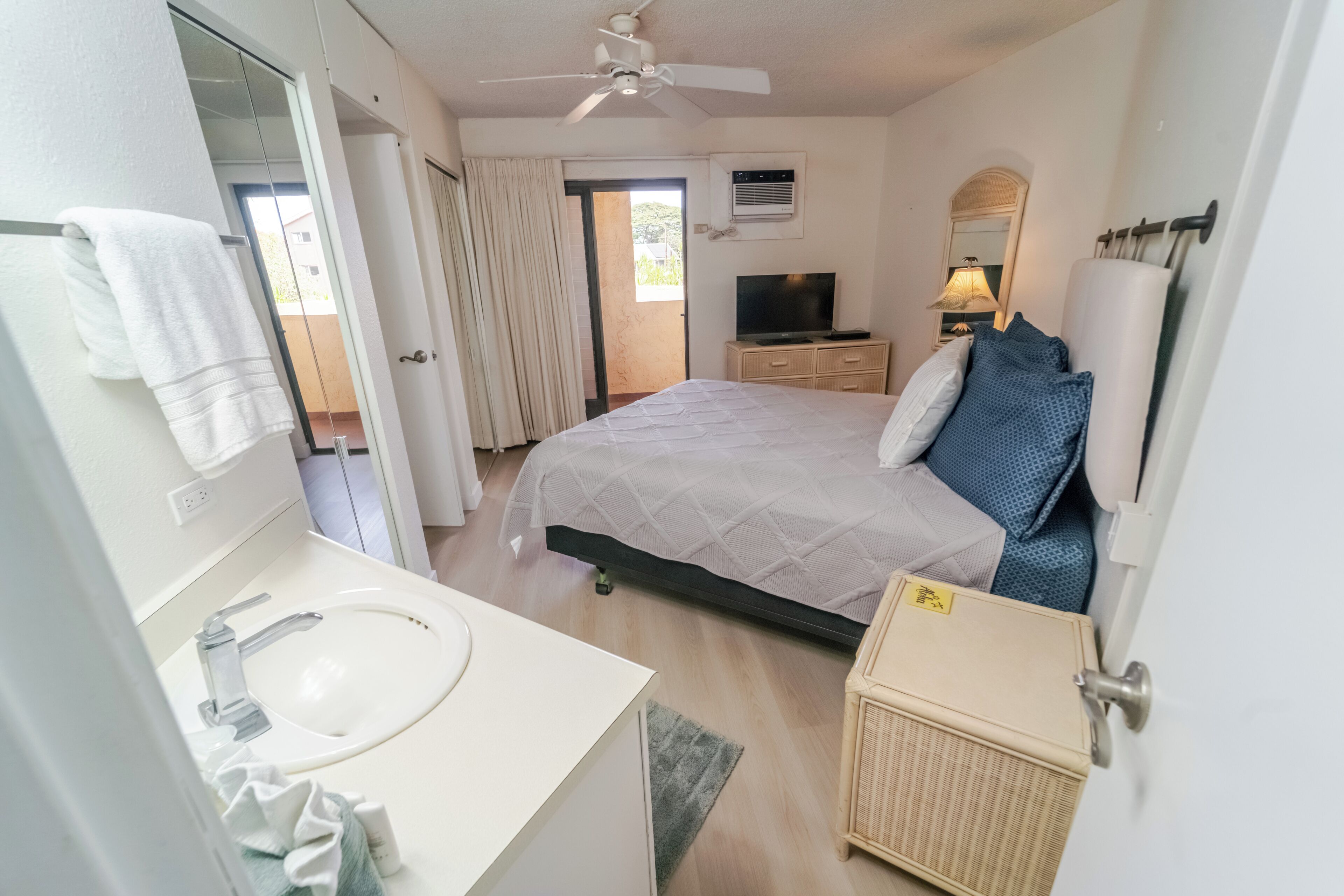 A bedroom with a bed, a TV, a bedside table, a mirror, and a bathroom with a sink and towel rack.