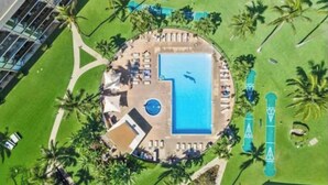 Eine Vogelperspektive auf einen Urlaubsort mit einem zentralen Swimmingpool, Liegebereichen und Palmen.