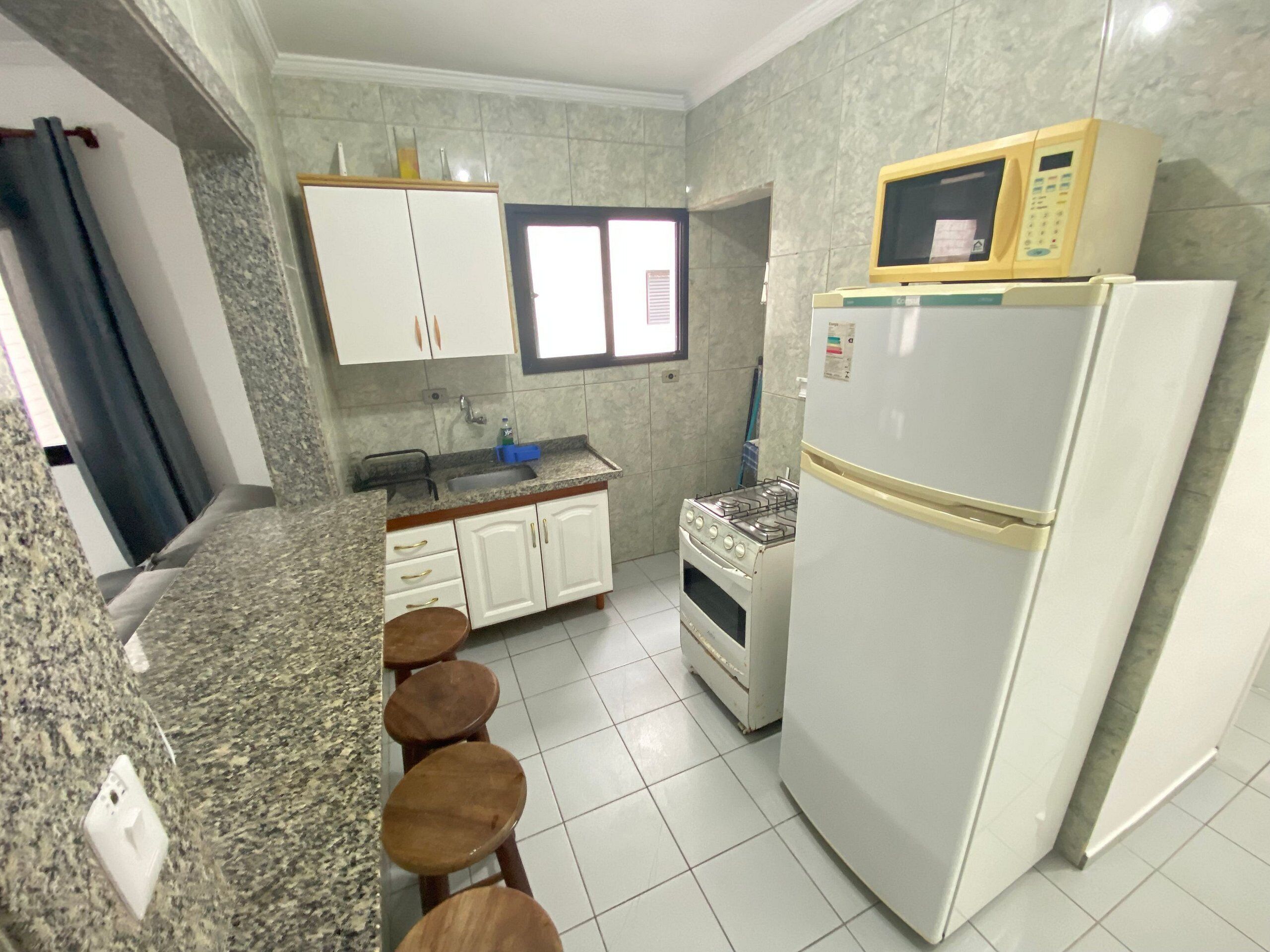 A kitchen with white cabinets, a stove, a microwave, and a refrigerator. There are stools at the counter and a window with blinds.