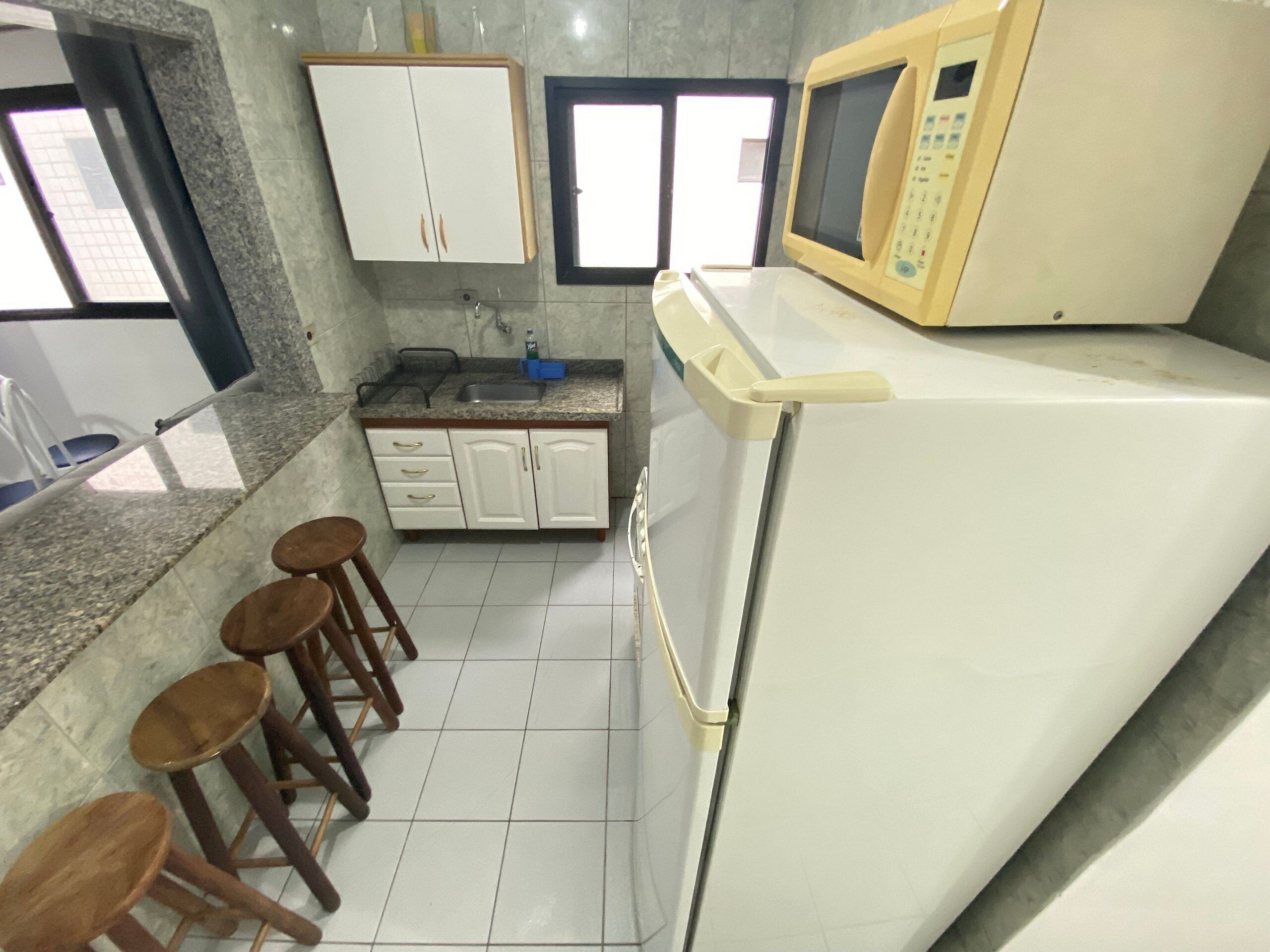A kitchen with white cabinets, a microwave, and a refrigerator. There are three wooden stools and a window with blinds.