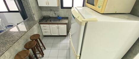 A kitchen with white cabinets, a microwave, and a refrigerator. There are three wooden stools and a window with blinds.