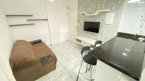 A compact living space with a sofa, a small dining area with a bar counter, a ceiling fan, and a flat-screen TV mounted on the wall.