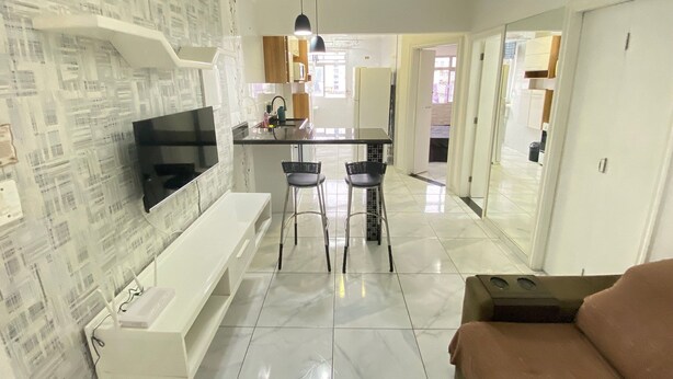 A modern living room with a flat-screen TV, a sofa, and a coffee table. There is a kitchen area with a dining table and chairs.