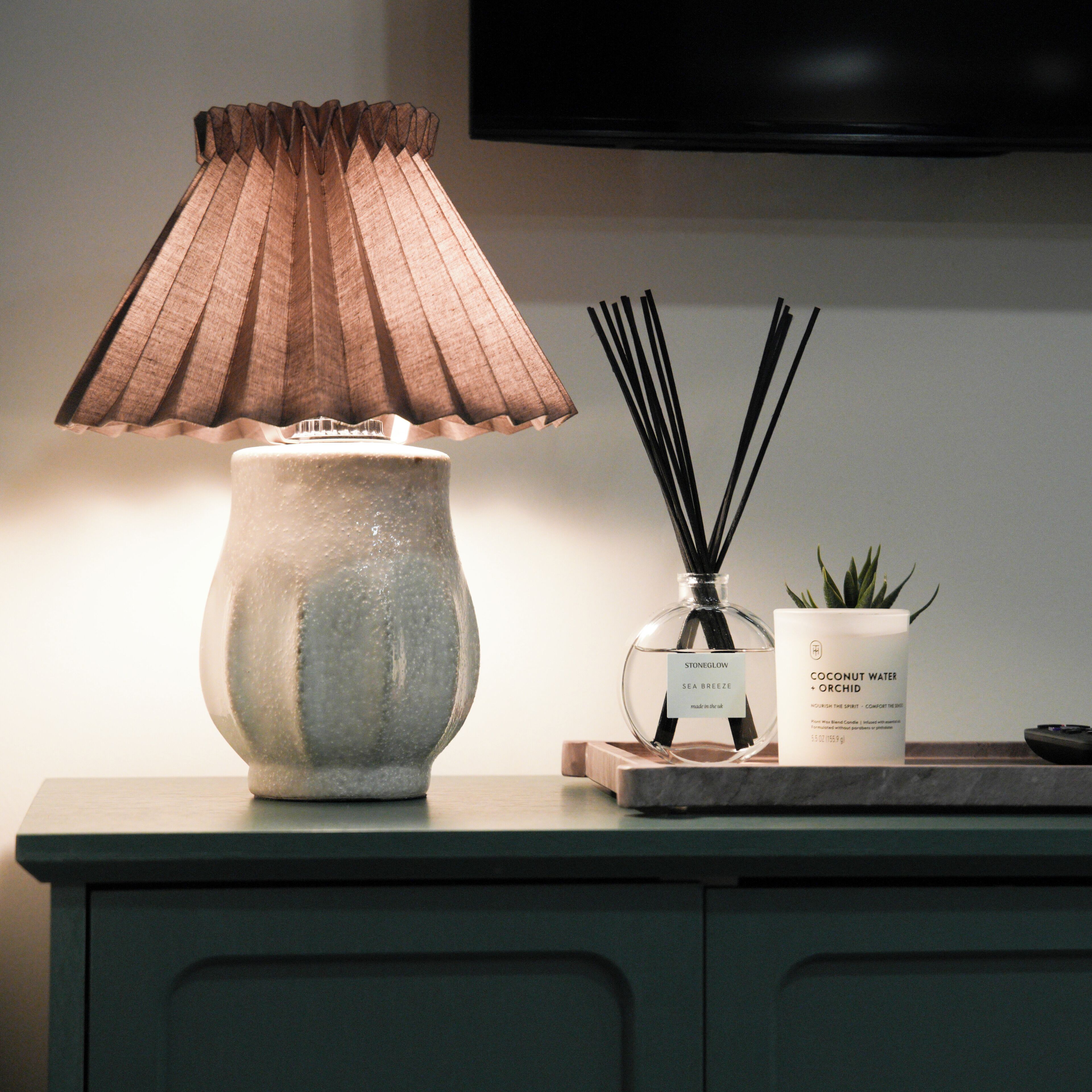 A lamp with a pleated shade, a bottle of reed diffuser, and a small potted plant on a wooden tray.