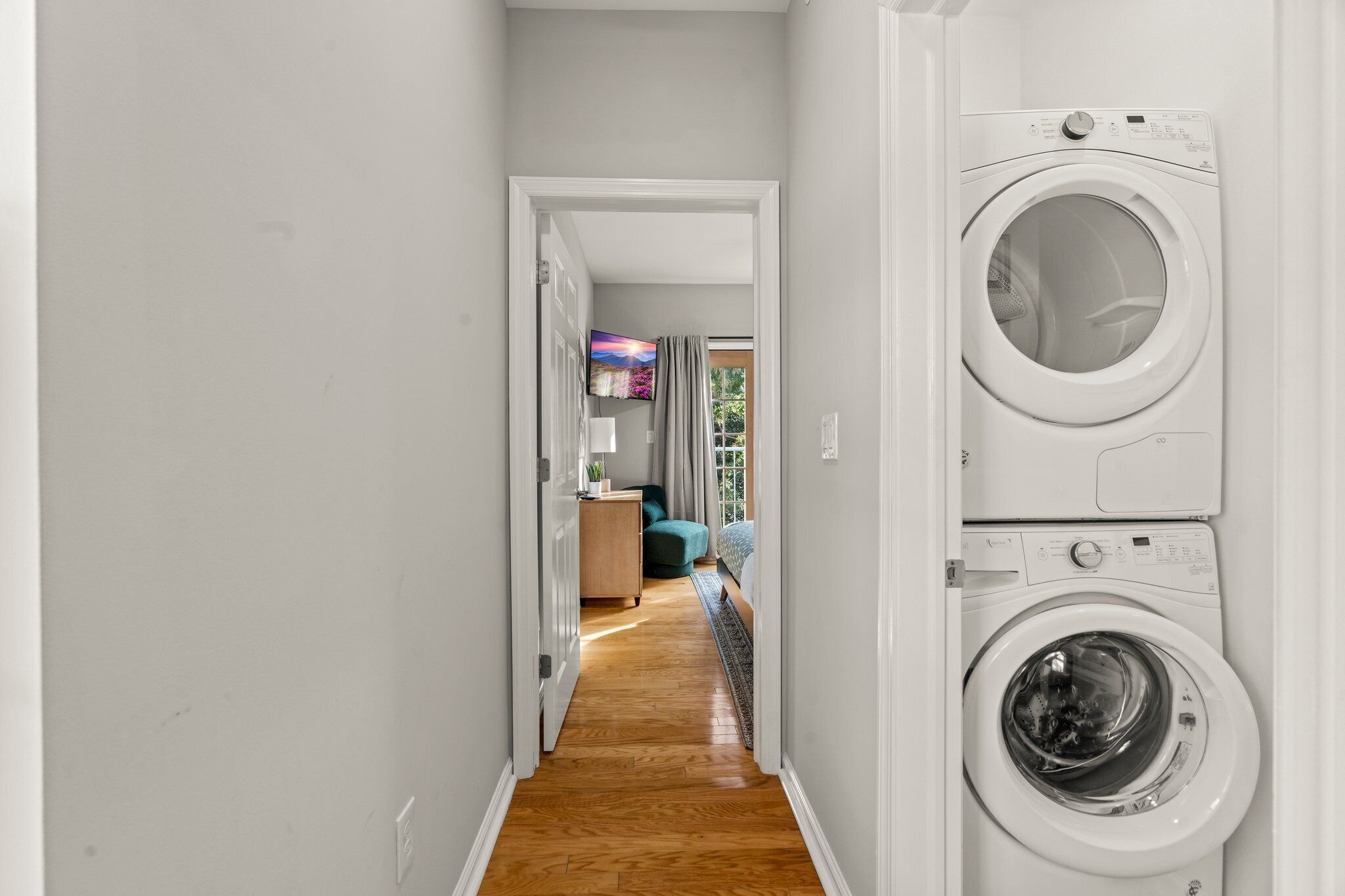 Une buanderie équipée d’un lave-linge et d’un sèche-linge, avec un plancher en bois, et un couloir menant à une chambre à coucher comprenant un lit et un téléviseur.