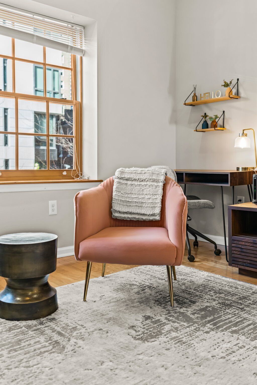 A modern room with a leather armchair, a round metal table, a patterned rug, a window with blinds, and a desk with shelves.