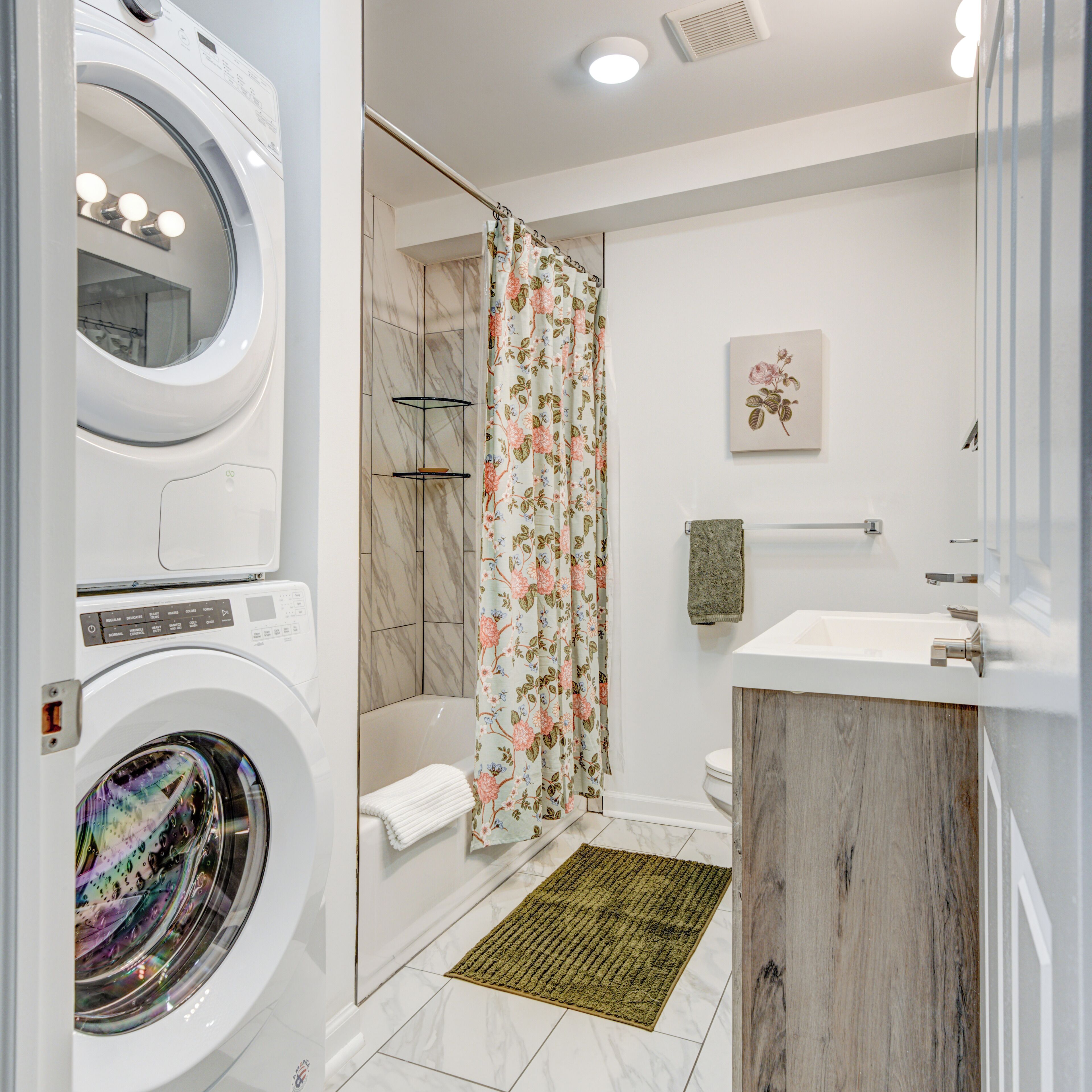 Een moderne badkamer met een ingebouwde wasmachine, een douche met een bloemen gordijn, een toilet en een wastafel met houten opbergmeubel.