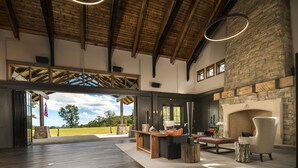 Un intérieur spacieux avec un plafond haut, des poutres apparentes, une cheminée en pierre et une vue sur l’extérieur.