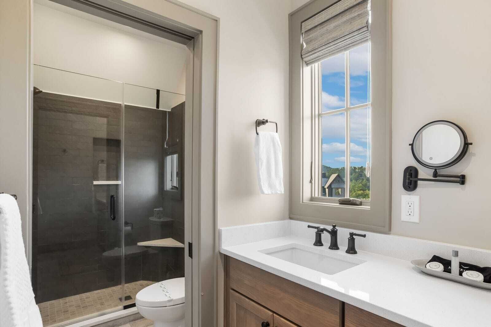 A modern bathroom with a glass-enclosed shower, a toilet, a sink with two faucets, a window with a view, and a round mirror on the wall.