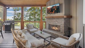Une terrasse extérieure avec des meubles en osier, une cheminée en pierre, un téléviseur fixé au-dessus de la cheminée et un barbecue.