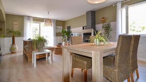 Ein Essbereich mit Holztisch und Korbstühlen, im Hintergrund eine Küche und üppige Kübelpflanzen.