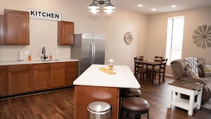 Una cocina con armarios de madera, un refrigerador de acero inoxidable, una isla con cubierta blanca y taburetes, y un comedor con mesa y sillas.