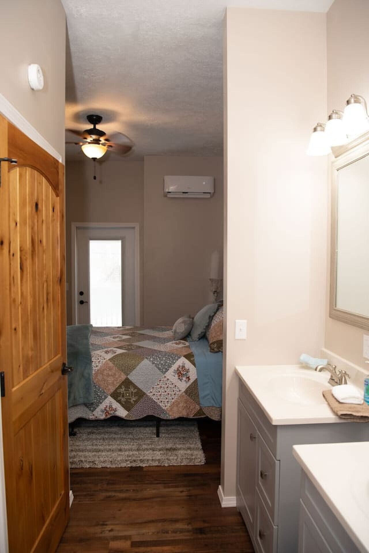A bedroom with a wooden door, a ceiling fan, a bed with a quilt, a bathroom with a sink and mirror, and a window.