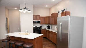 Una cocina moderna con armarios de madera, un refrigerador de acero inoxidable, una isla central con taburetes y un letrero que indica el área de cocina.