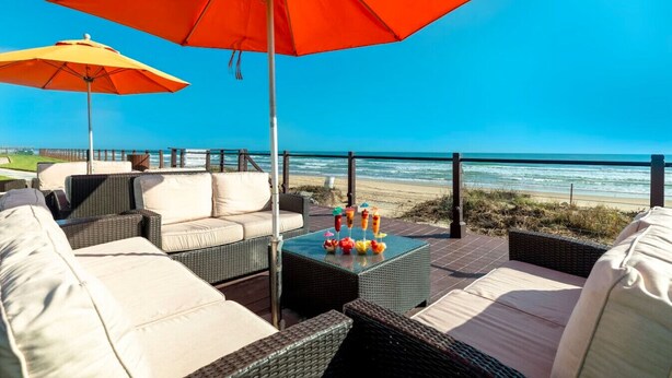 Une terrasse au bord de la plage, avec des meubles en osier, une table en verre sur laquelle sont posées des boissons, et deux grands parasols.