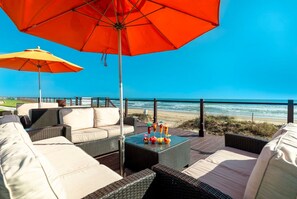 Une terrasse au bord de la plage, avec des meubles en osier, une table en verre sur laquelle sont posées des boissons, et deux grands parasols.