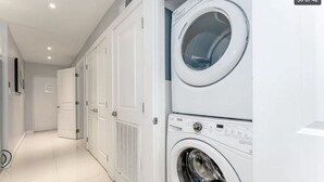 Une buanderie équipée d’une machine à laver et d’un sèche-linge superposés, avec un sol carrelé et une porte donnant sur une autre pièce.
