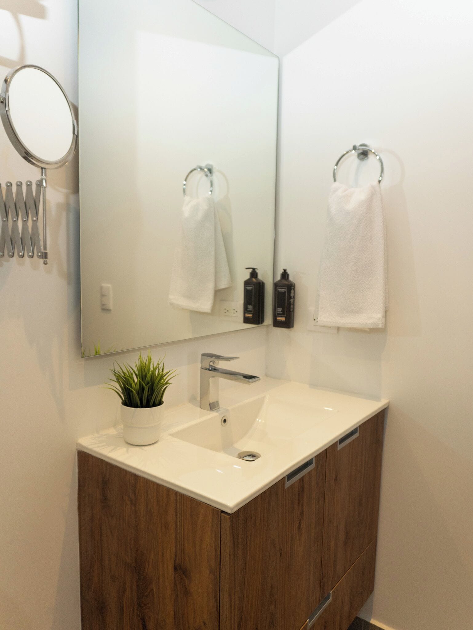Ein modernes Badezimmer mit einem weißen Waschbecken, einem Holfach und einem großen Spiegel. Über dem Spiegel hängen zwei weiße Handtücher an Handtuchhaltern, und auf dem Waschtisch steht ein kleiner Topfpflanze.