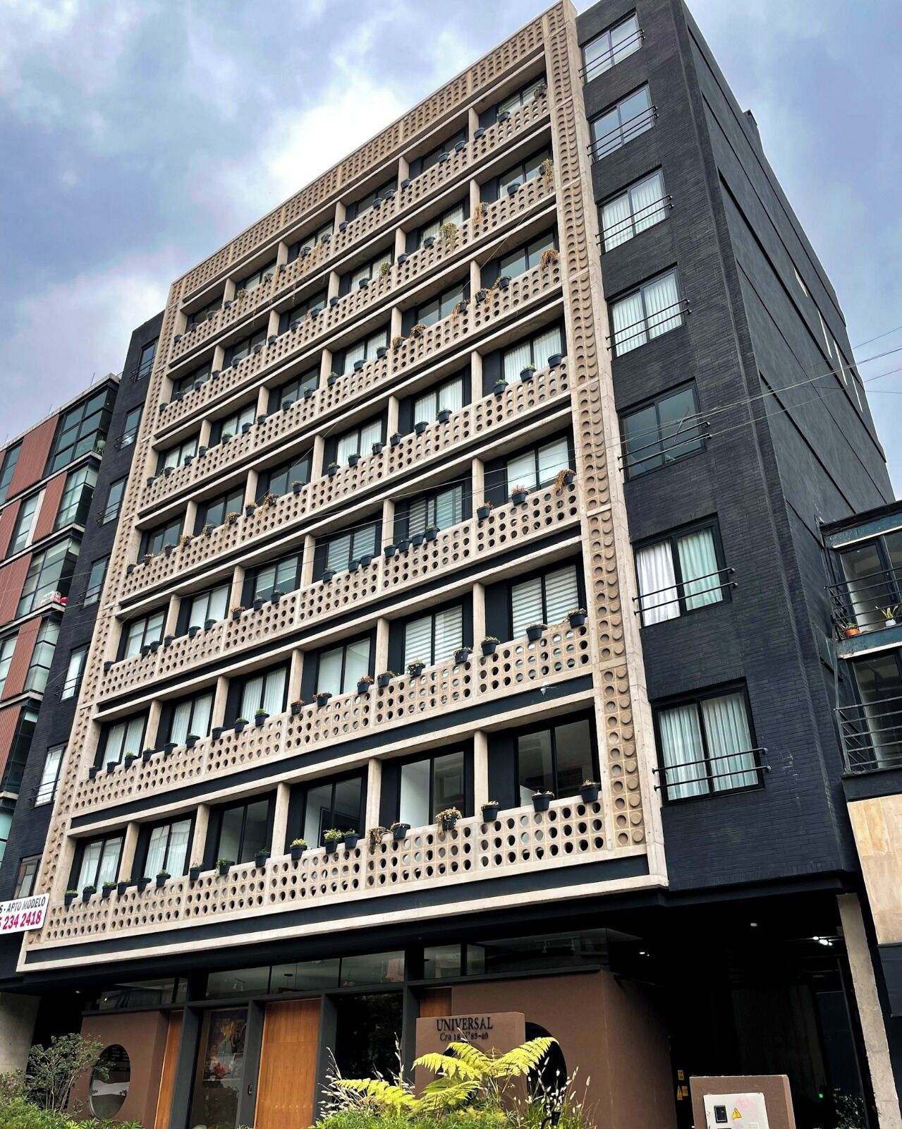 Un edificio de varios pisos con un diseño arquitectónico moderno, que cuenta con balcones con plantas en macetas y un letrero que indica que es un hotel.