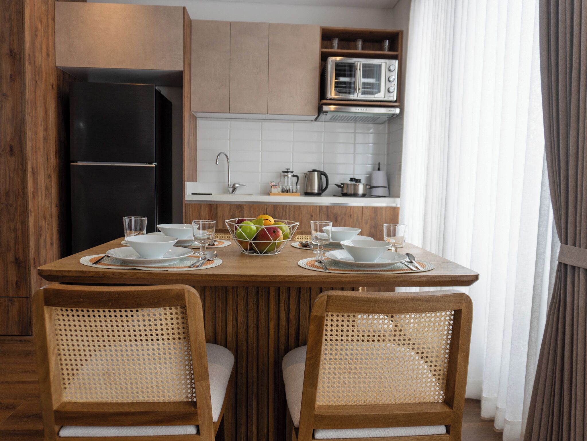 A modern kitchen with wooden cabinets, a black refrigerator, a microwave, and a dining table set with white plates, bowls, and a fruit bowl.