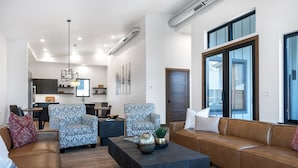 A modern living room with a brown leather sofa, patterned armchairs, a dark coffee table, and a large window with blinds.