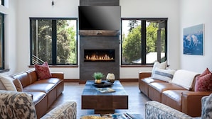 A modern living room with a fireplace, leather sofas, and large windows offering a view of trees.