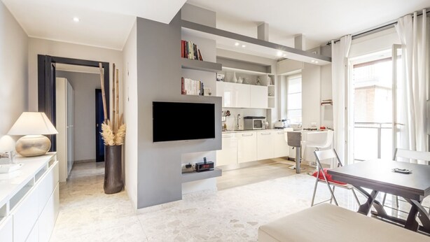 Un salon moderne doté d’un téléviseur à écran plat, d’un coin repas avec une table et des chaises, et d’une cuisine avec des meubles blancs et un four à micro-ondes.