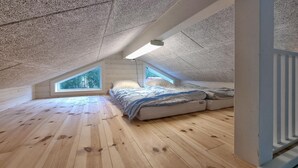 Ein Schlafzimmer im Loft-Stil mit schrägverdeckter Decke, Holzboden, einem Bett mit Matratze und Kissen sowie zwei Oberlichtern, die für natürliches Licht sorgen.