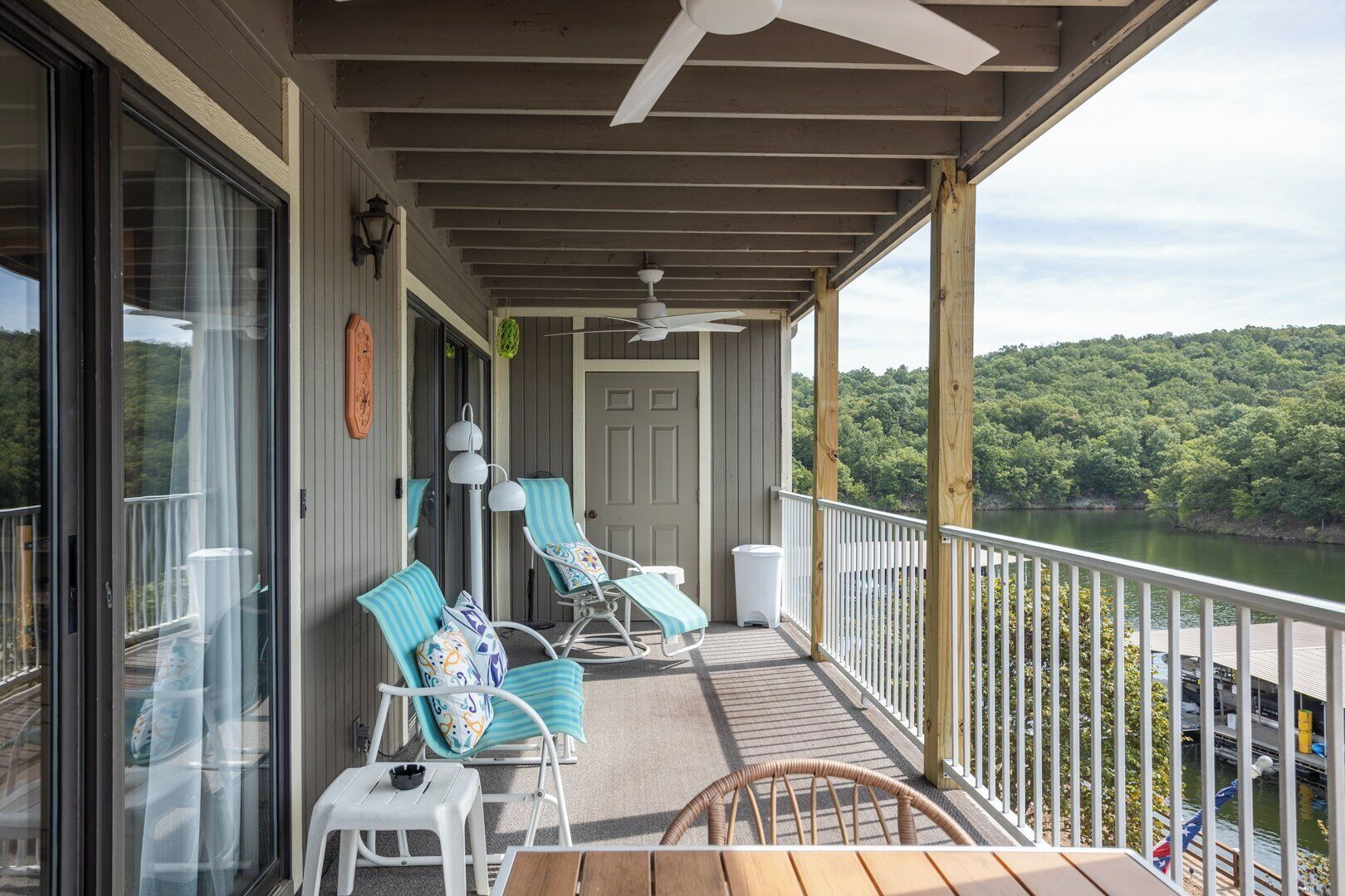 A covered outdoor deck with lounge chairs and a table, overlooking a body of water and forested hills.