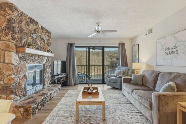 Ein Wohnzimmer mit Kamin aus Stein, Deckenventilator, einer Couch, einem Couchtisch und einem Balkon mit Landschaftsblick.