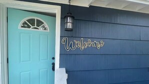 A turquoise door with a white trim and a decorative window above it. A 'Welcome' sign is mounted on the wall to the right of the door.