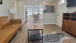 A spacious living room with a leather sofa, a coffee table, and a zebra rug. There is a TV mounted on the wall, a wooden cabinet, and a dining area with chairs and a plant.