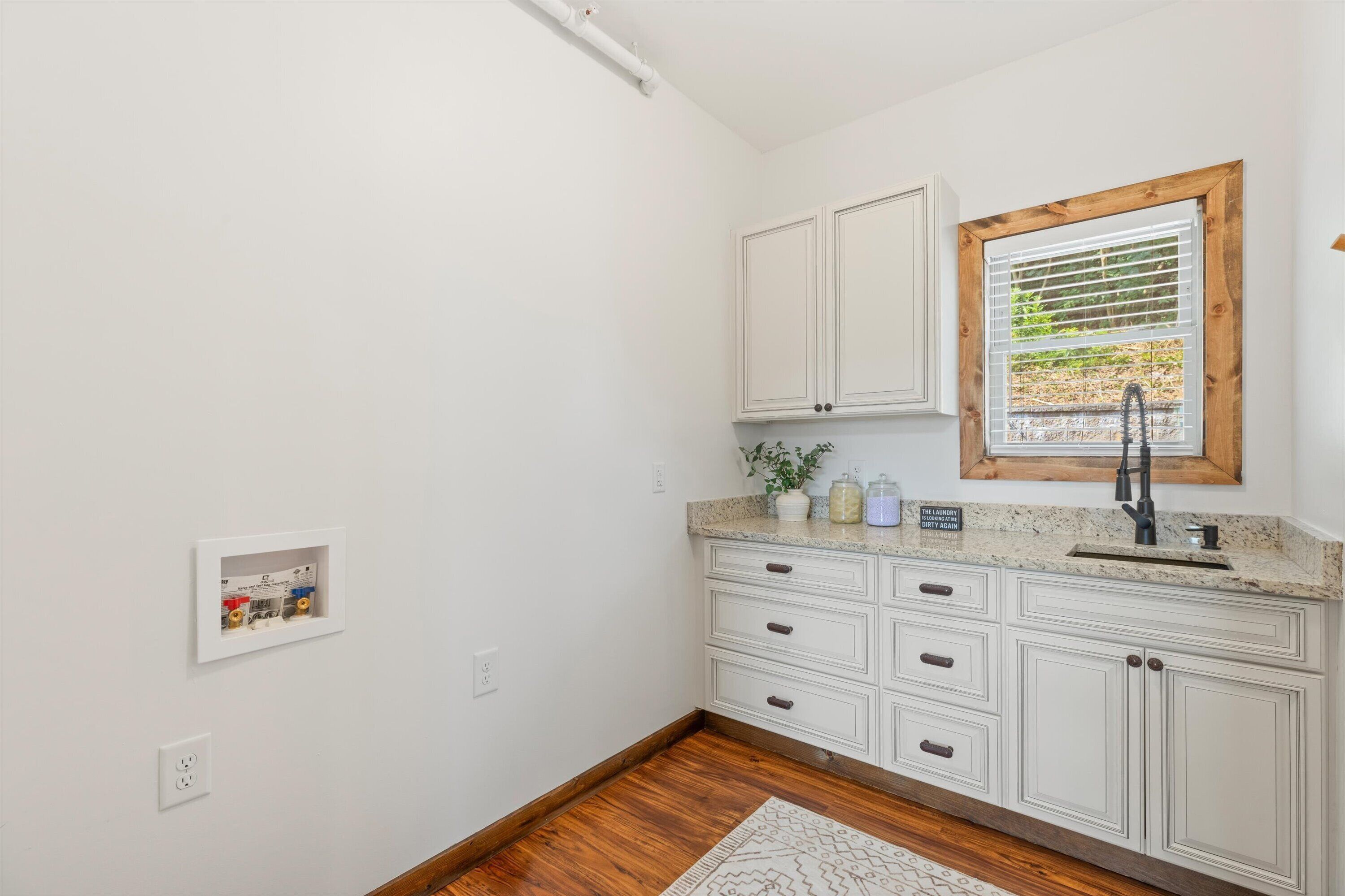 A kitchen with white cabinets, a granite countertop, a window with blinds, and a wooden floor.