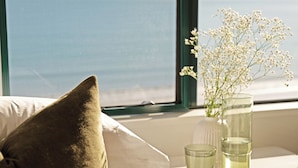 Una cama con un almohadón, una mesita de noche con un florero y vistas al mar a través de una ventana.