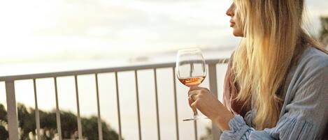 Eine Person sitzt auf einem Balkon, an einem gedeckten Tisch, mit einem Weinglas in der Hand und blickt auf eine schöne Aussicht.