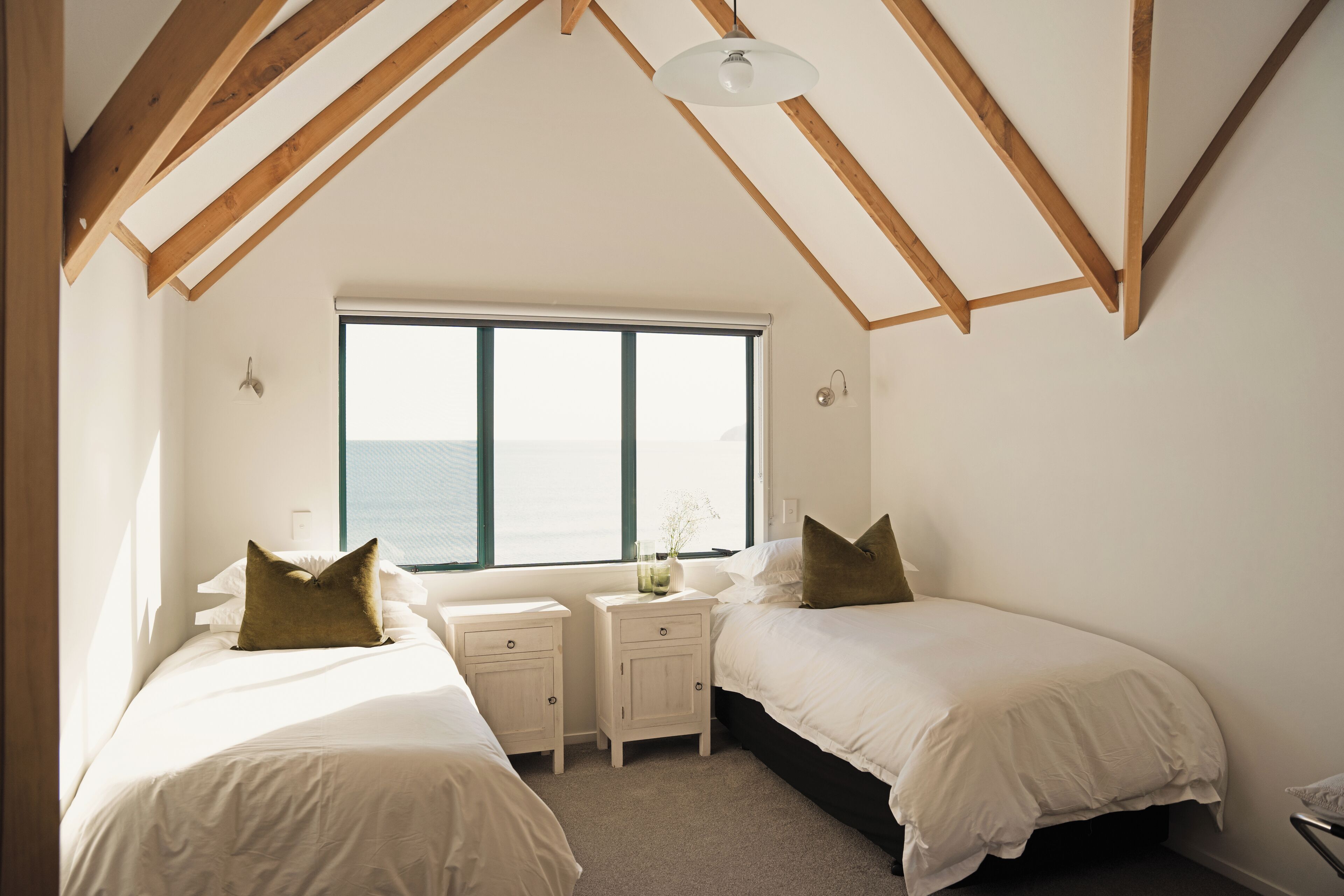 A bedroom with two beds, a large window, wooden beams, and a small bedside table with a lamp.