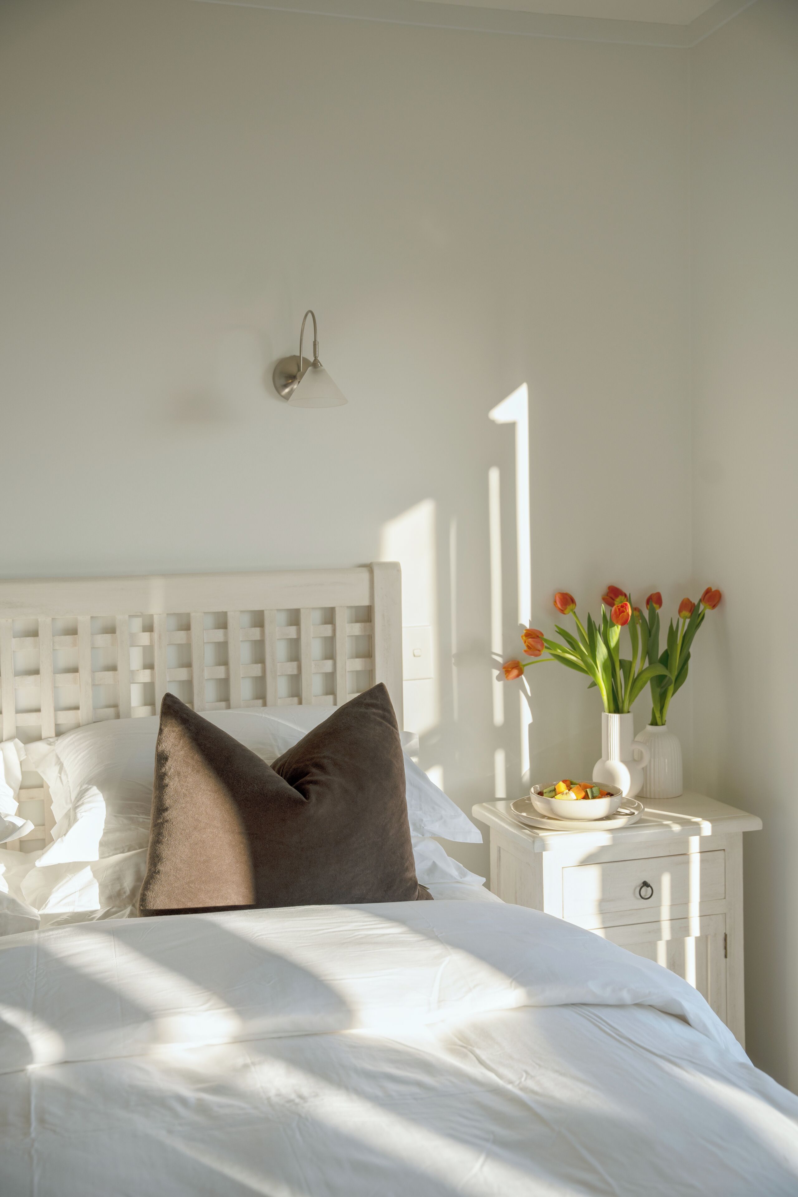 Una cama bien hecha con almohadas, una mesita de noche con un florero y una lámpara de pared.