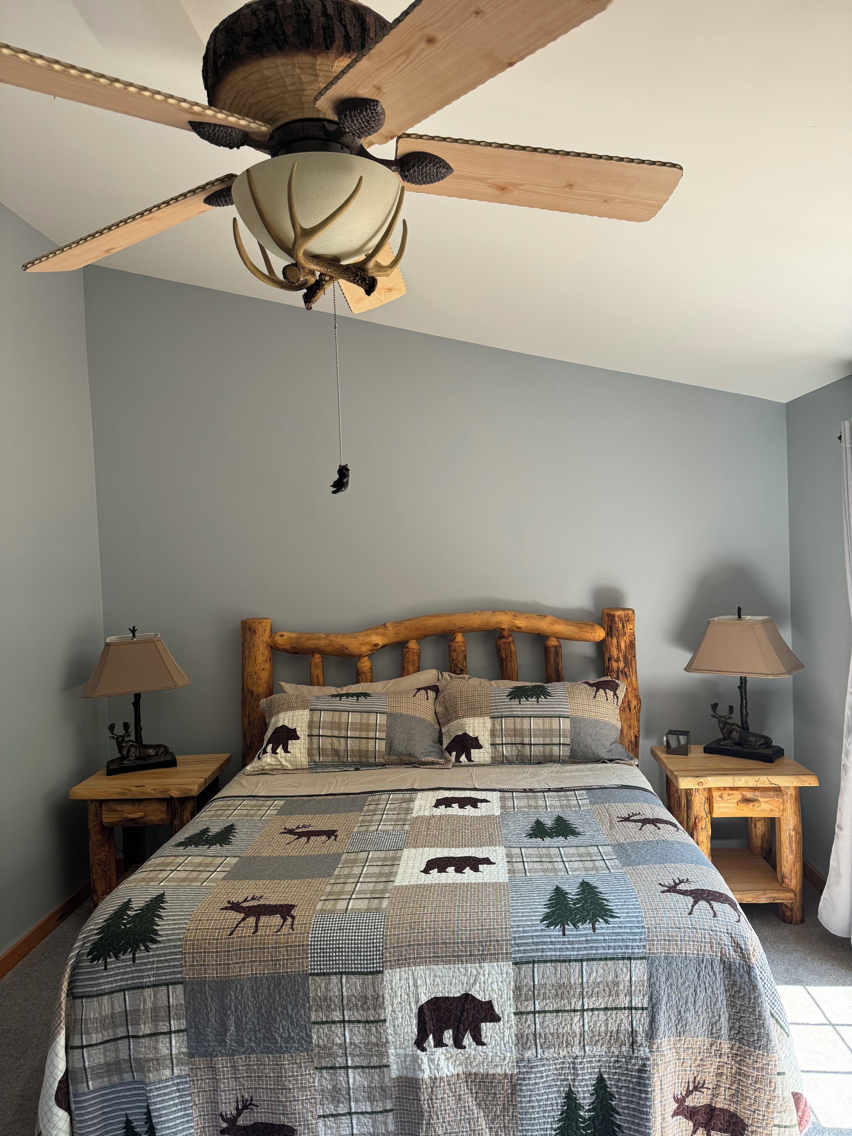 A bedroom with a wooden headboard, a ceiling fan, a quilt with animal prints, two bedside tables with lamps, and a window with curtains.