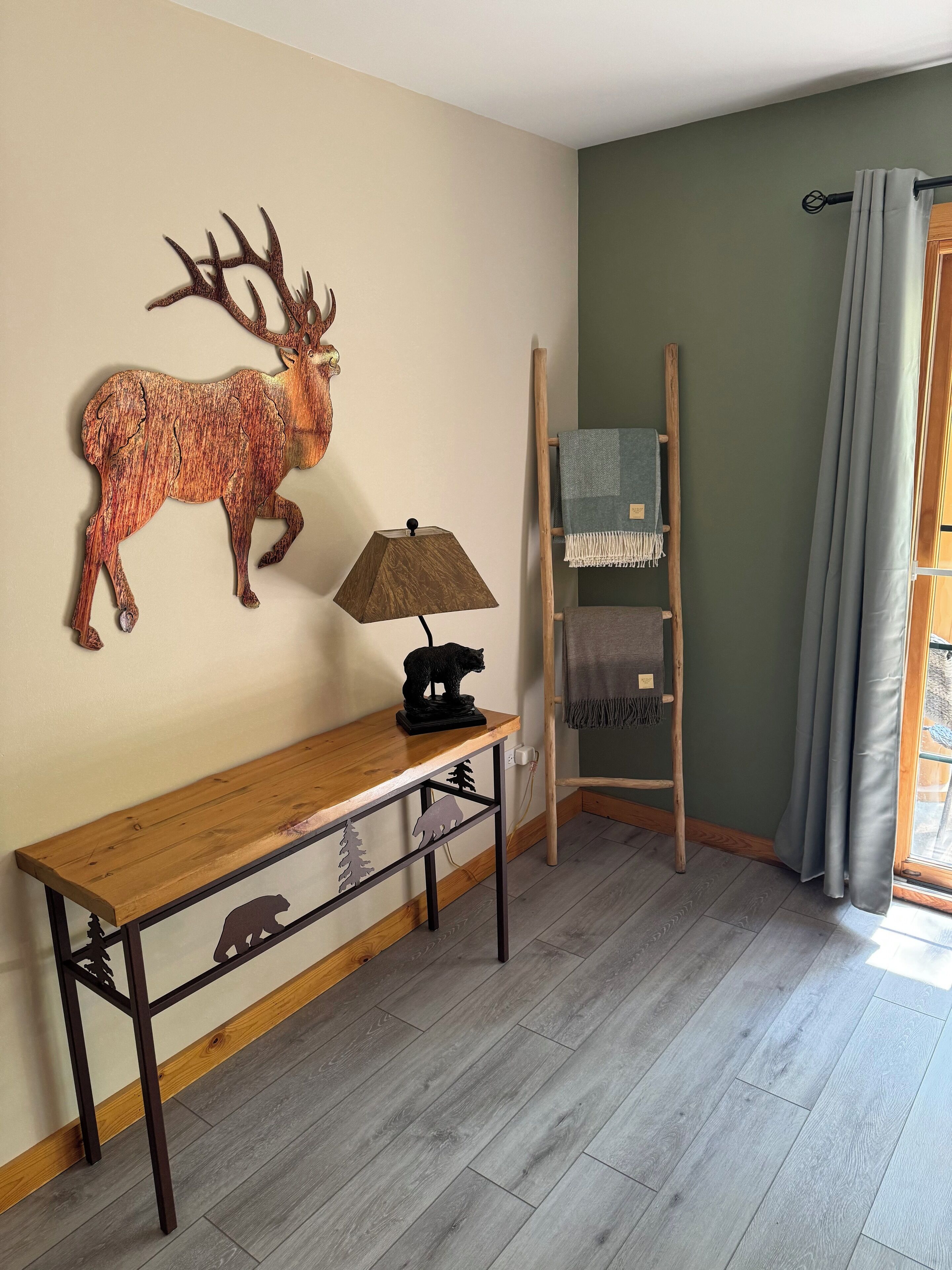 Una habitación con un banco de madera, una escultura de ciervo montada en la pared, una lámpara, una escalera y una ventana con cortinas.