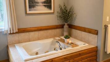 A bathroom with a large wooden bathtub, a framed landscape painting, and a potted plant.