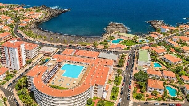 Luftaufnahme eines Küstenresorts mit einem zentralen Schwimmbecken, mehreren Gebäuden mit roten Dächern und einer felsigen Küstenlinie.