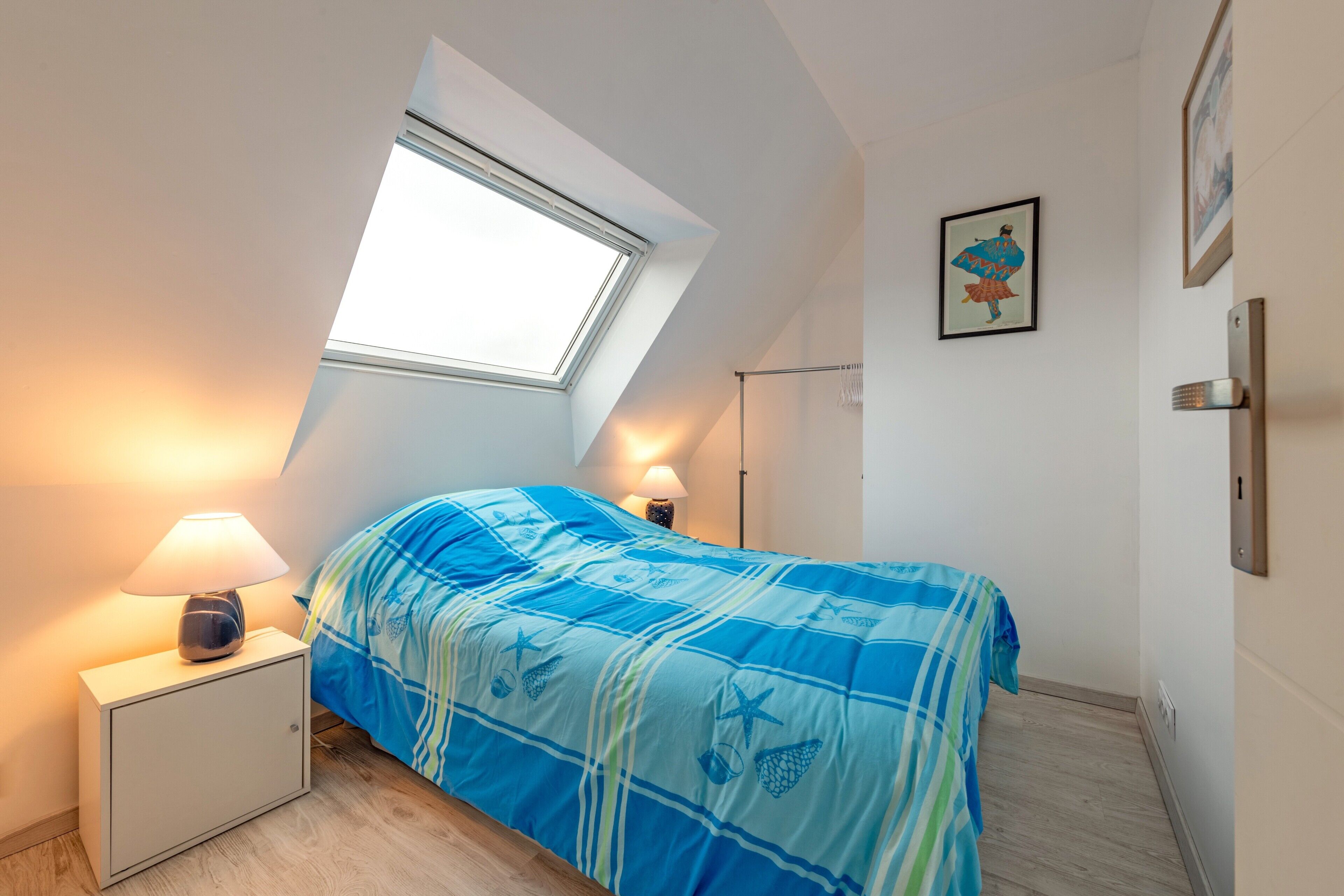 A bedroom with a skylight, a bed with a blue and green patterned bedspread, a bedside lamp, and a framed picture on the wall.